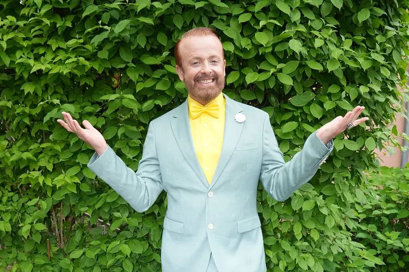 Ross Antony vor einem grünen Busch in einem blauen Anzug mit gelbem Hemd