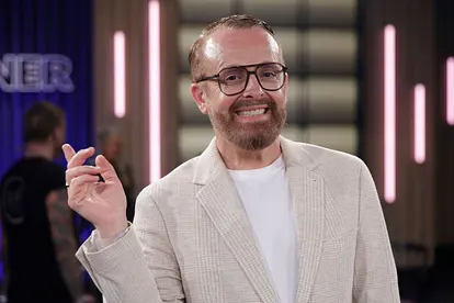 Ross Antony, hier mit Brille, weißem T-Shirt und beigem Cord-Blazer zu sehen, hebt eine Hand und lächelt in die Kamera.