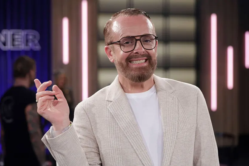 Ross Antony, hier mit Brille, weißem T-Shirt und beigem Cord-Blazer zu sehen, hebt eine Hand und lächelt in die Kamera.