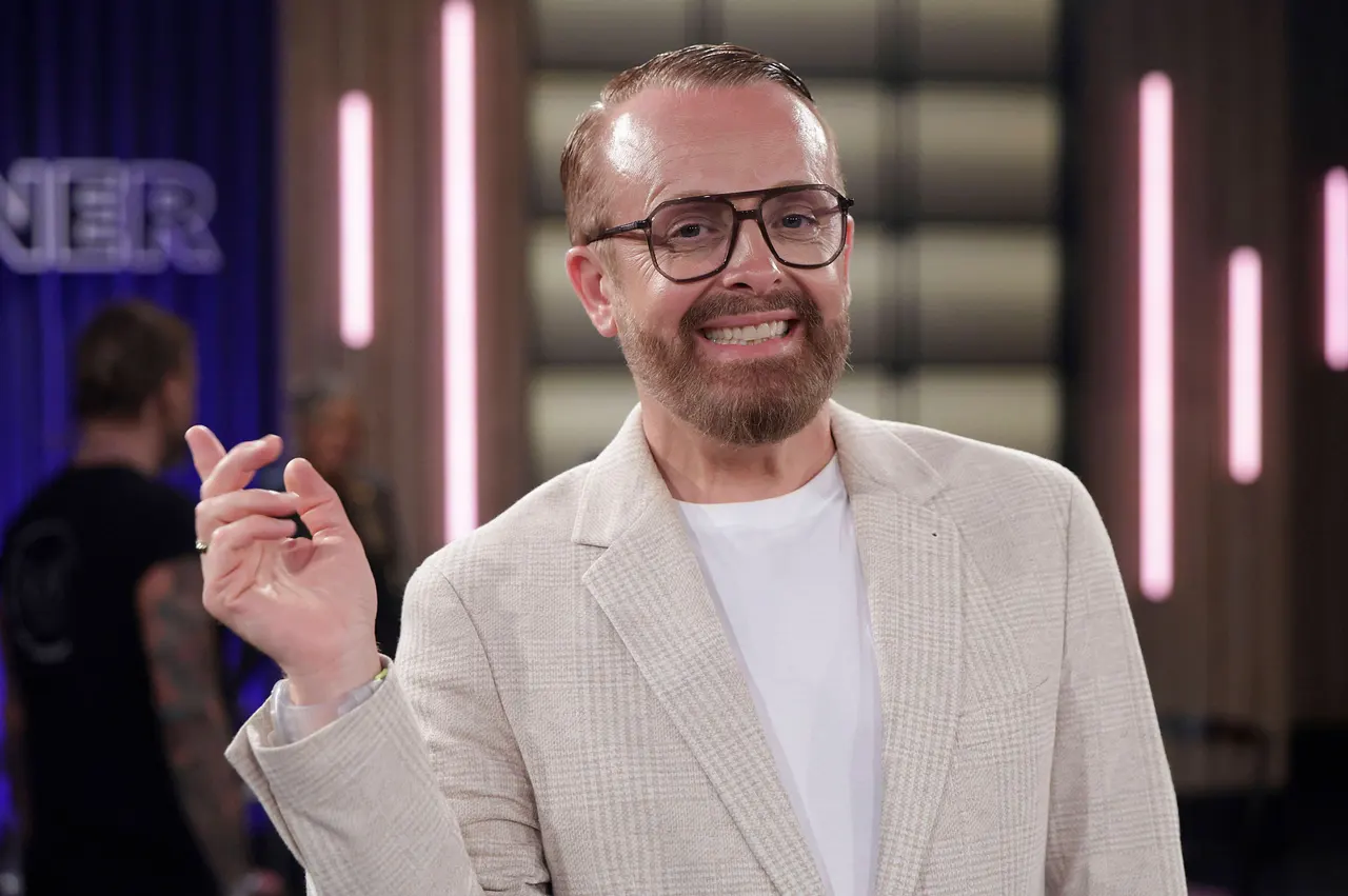 Ross Antony, hier mit Brille, weißem T-Shirt und beigem Cord-Blazer zu sehen, hebt eine Hand und lächelt in die Kamera.