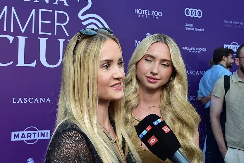 Shania und Davina Geiss stehen vor einer Fotowand des Lascana Summer Club-Events. Ihnen wird ein Mikrofon entgegengehalten. Im Hintergrund sieht man zwei Männer.