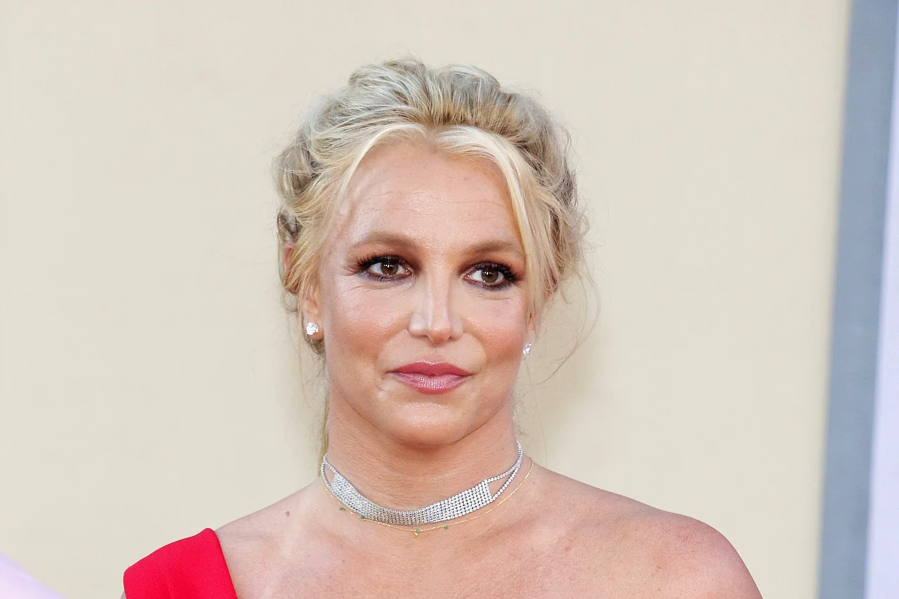 Britney Spears trägt eine wuschelige Hochsteckfrisur und ein rotes Kleid mit nur einem Träger. Sie lächelt sanft. 