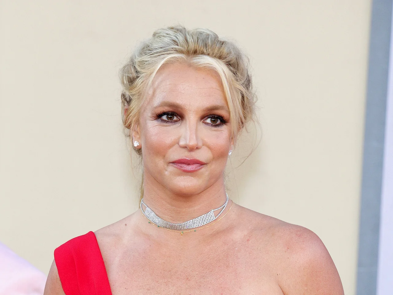 Britney Spears trägt eine wuschelige Hochsteckfrisur und ein rotes Kleid mit nur einem Träger. Sie lächelt sanft.  - Foto: IMAGO / Depositphotos