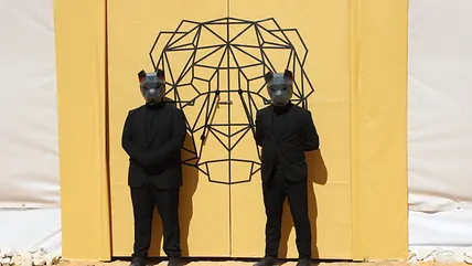 Zwei Personen in schwarzer Kleidung und mit schwarzen Löwenmasken stehen vor zwei gelben Türen, auf denen ein stilisierter Löwenkopf zu sehen ist.