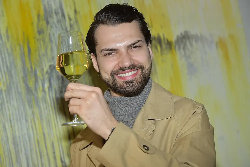 Jimi Blue Ochsenknecht trägt einen beigen Trenchcoat und hält grinsend ein Glas Weißwein hoch. Jimi Blue Ochsenknecht trägt einen beigen Trenchcoat und hält grinsend ein Glas Weißwein hoch.