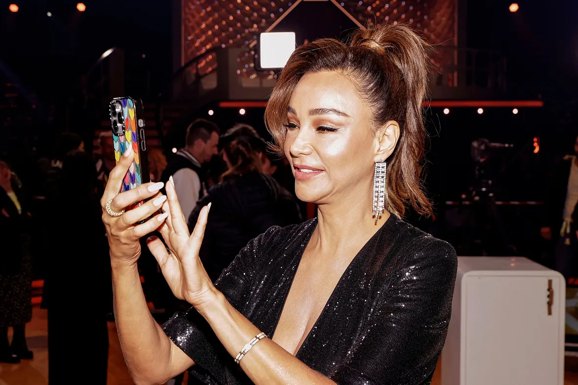 Verona Pooth, in einem Glitzerkleid und mit hochgebundenen Haaren, tippt etwas auf ihrem Handy ein.