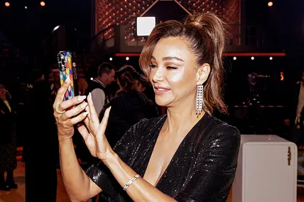 Verona Pooth, in einem Glitzerkleid und mit hochgebundenen Haaren, tippt etwas auf ihrem Handy ein.