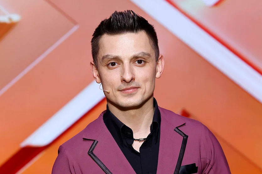 Zsolt Sándor Cseke steht vor einem orangenem Hintergrund und grinst in die Kamera. 
