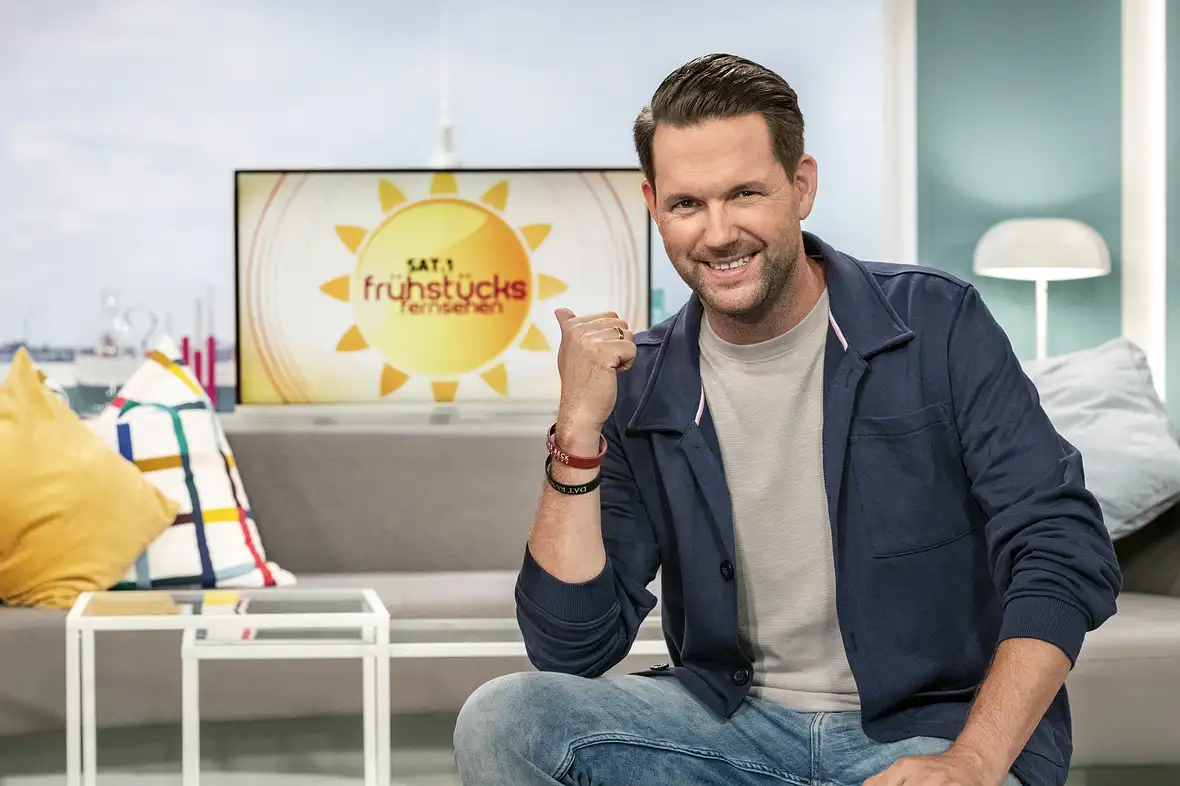 Matthias Killing posiert sitzend und lächelnd vor dem "Frühstücksfernsehen"-Logo
