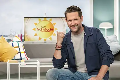 Matthias Killing posiert sitzend und lächelnd vor dem "Frühstücksfernsehen"-Logo