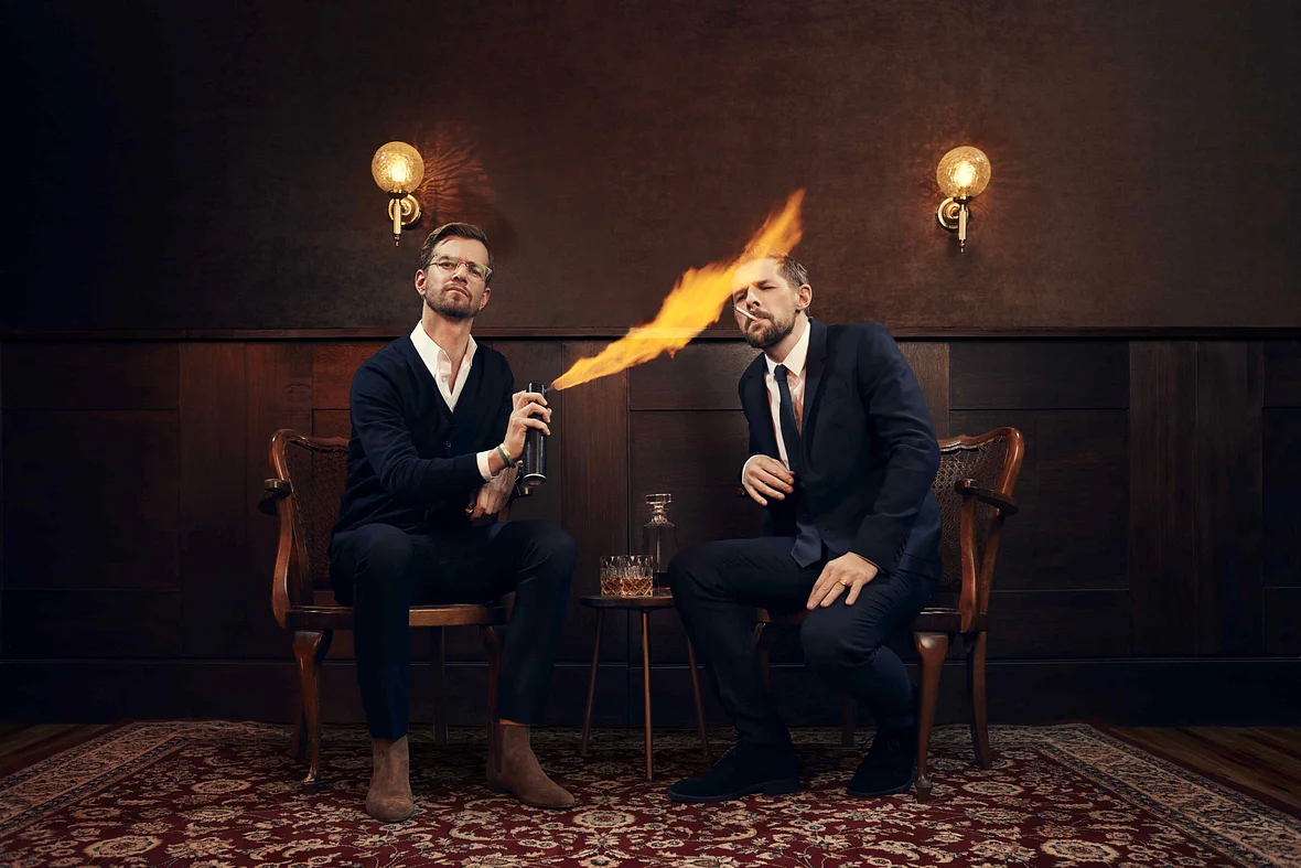Joko und Klaas sitzen auf Holzstühlen. Joko zündet Klaas' Zigarette mit einer brennenden Spraydose an.