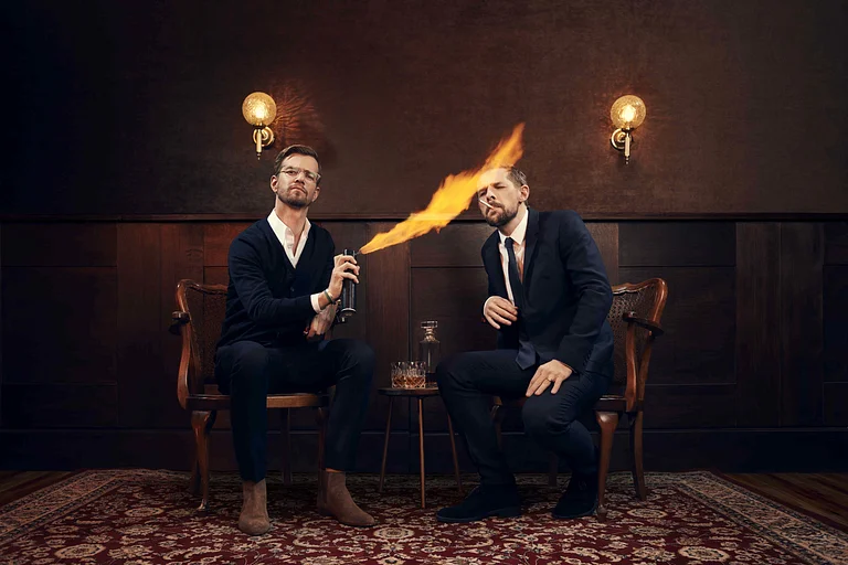 Joko und Klaas sitzen auf Holzstühlen. Joko zündet Klaas' Zigarette mit einer brennenden Spraydose an.